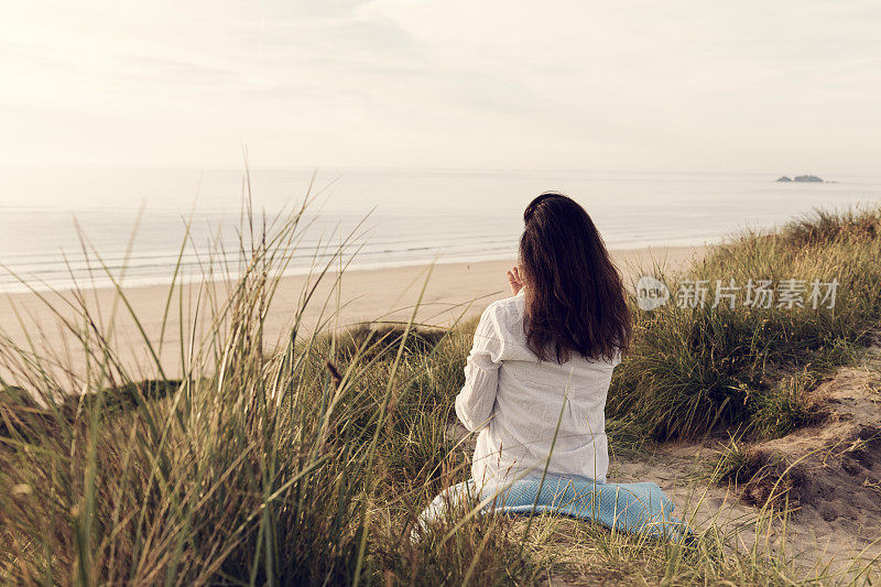 一名女子坐在沙丘上，望着日落时分康沃尔郡的Gwithian Towans海滩。